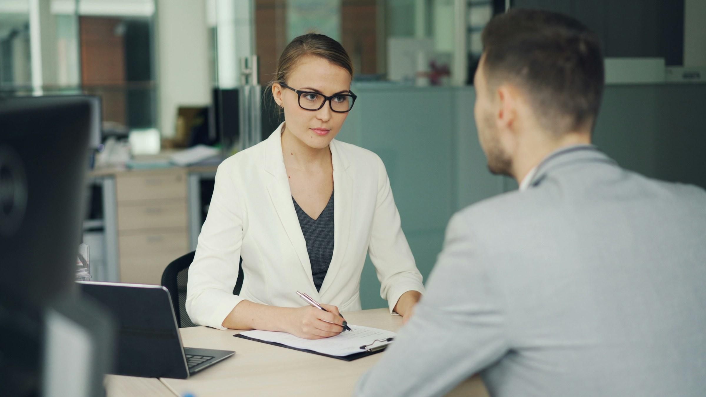 A recruiter in a white blazer interviewing a candidate across an office desk
